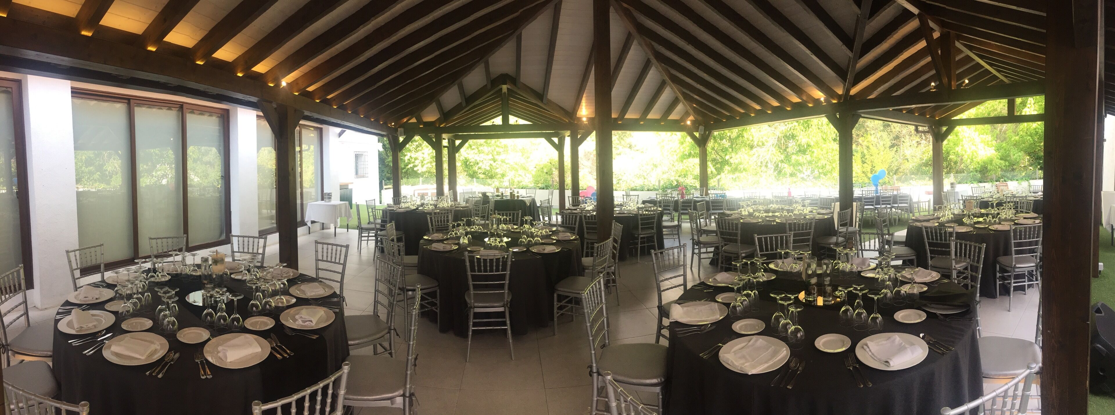An outdoor dining area with multiple round tables set for a formal event, surrounded by chairs and covered by a wooden pavilion.