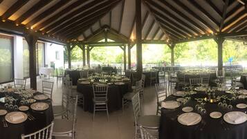 An outdoor dining area with multiple round tables set for a formal event, surrounded by chairs and covered by a wooden pavilion.