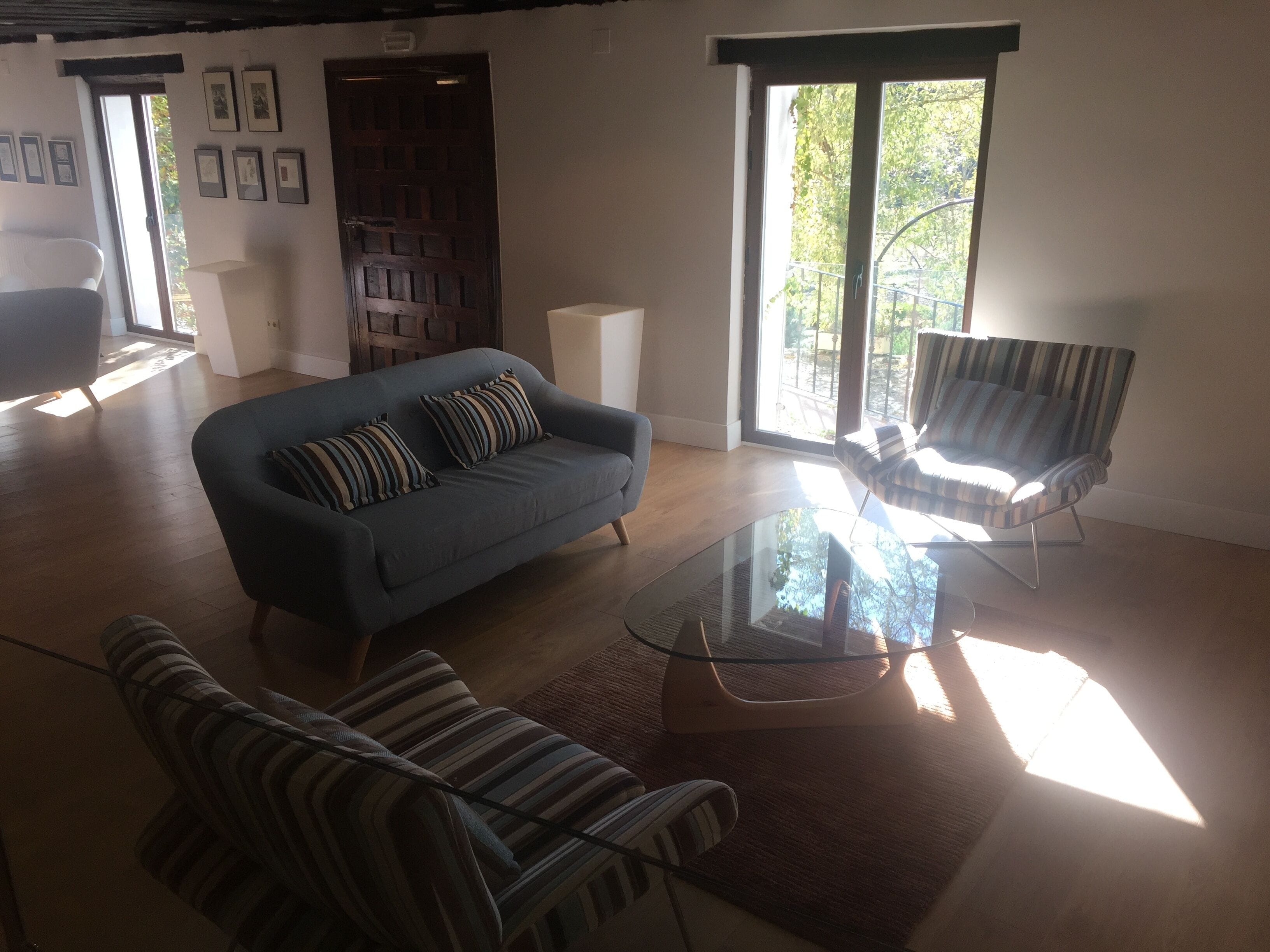 A spacious living room with a blue sofa, striped armchairs, a glass coffee table, and large windows offering a view of greenery outside.