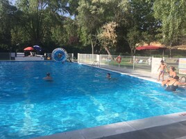 Una piscina con agua azul cristalina, rodeada de árboles y una zona vallada con sillas de descanso y sombrillas.