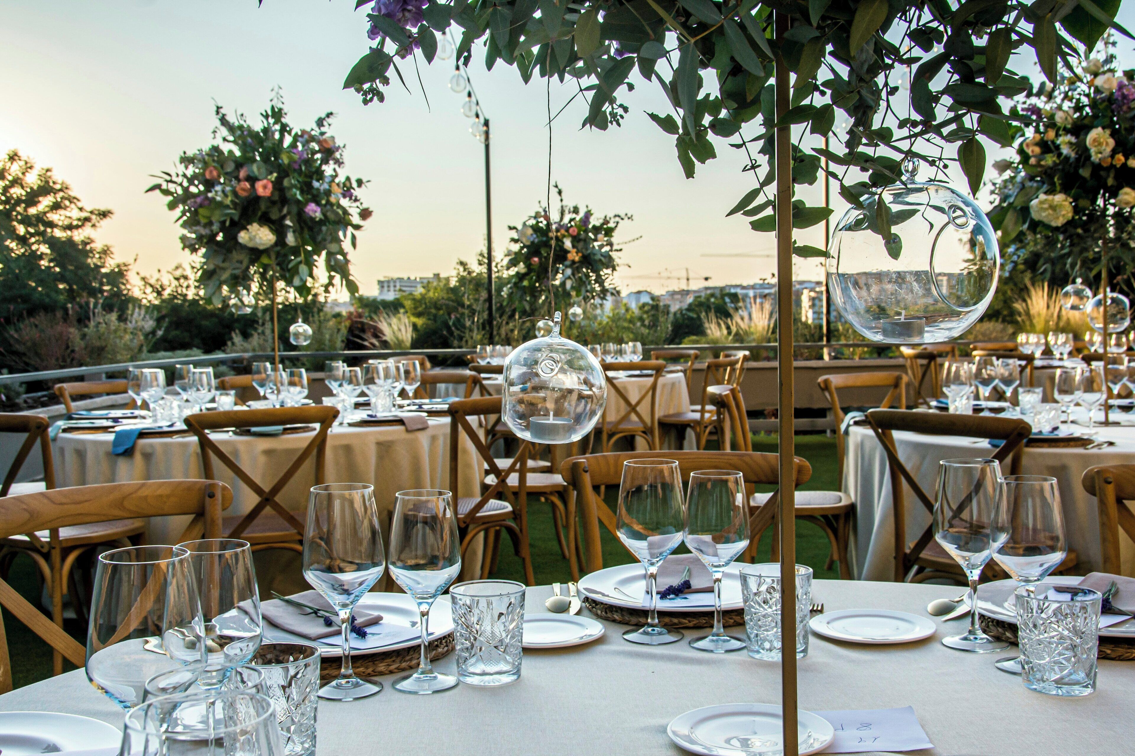 Un espacio de comedor al aire libre con mesas redondas, sillas y vasos de vidrio transparente.