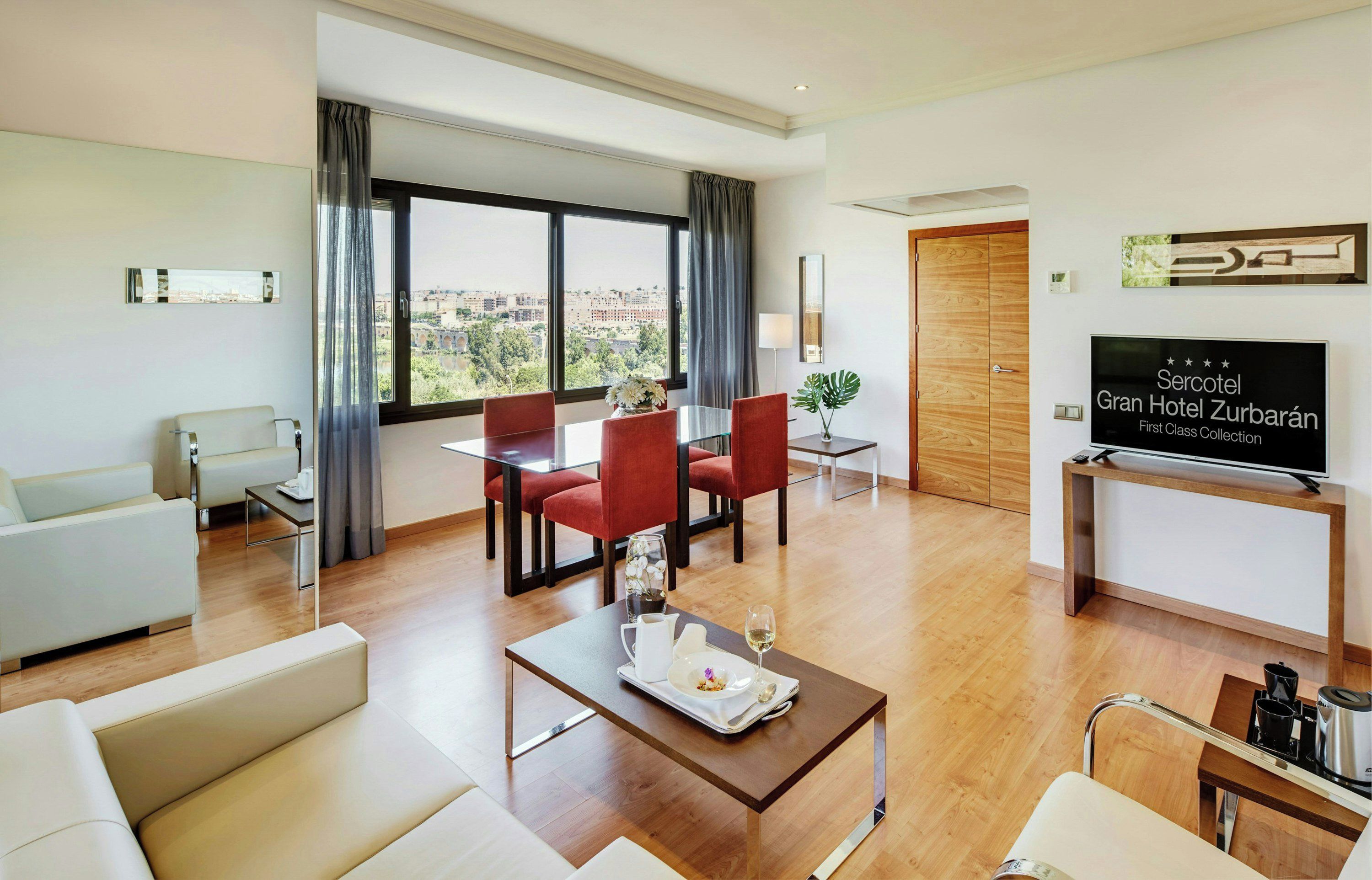 A hotel room with a dining area, a sofa, a TV, and a view of the city.