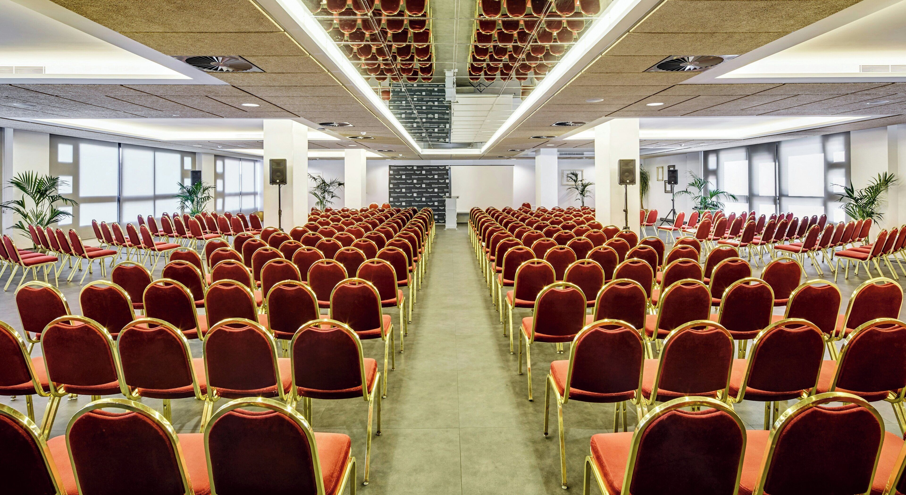 A conference room with rows of red chairs and gold frames, large windows, and a central stage.