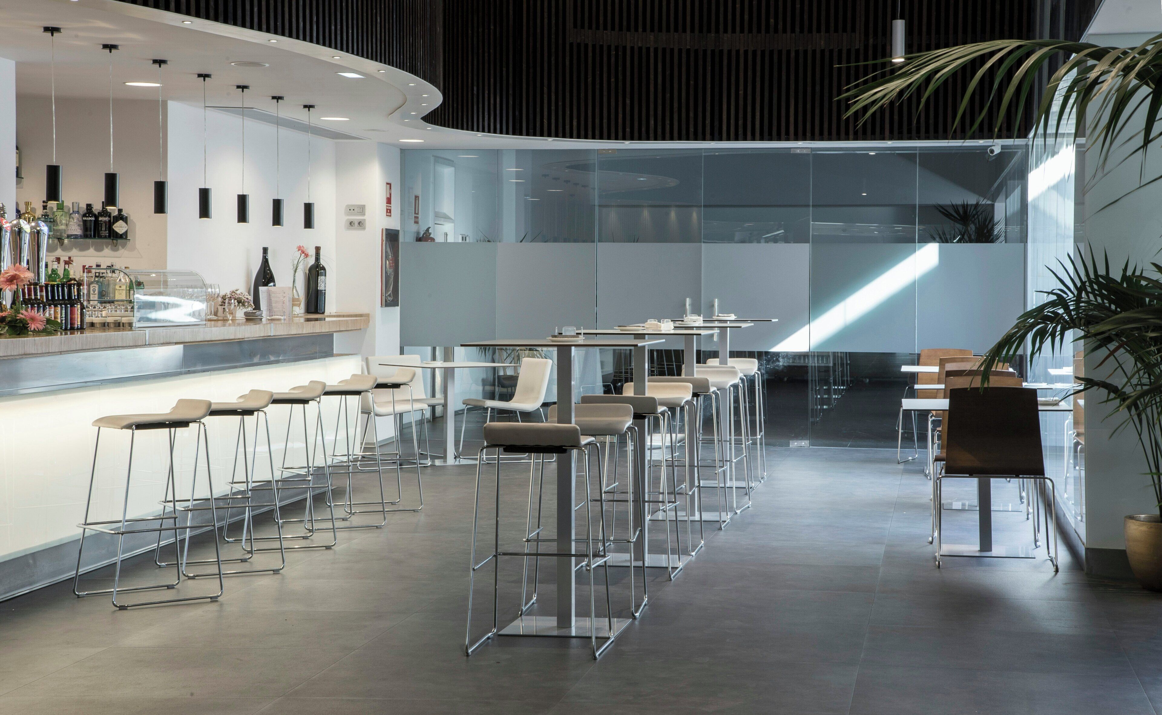 Zona de bar moderna con taburetes altos, una barra repleta de botellas y un separador de vidrio.
