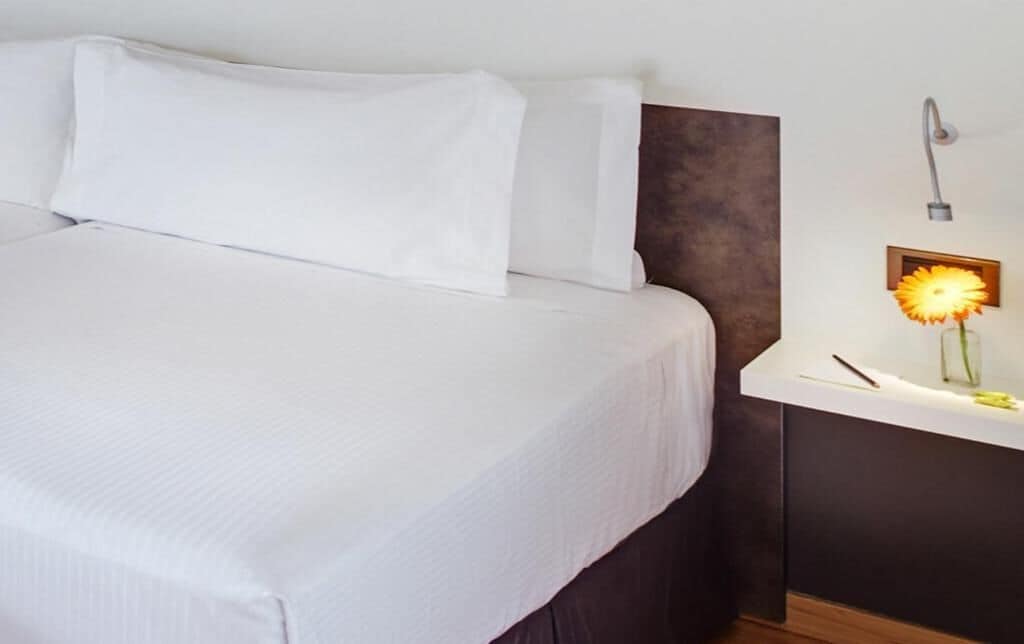 Una cama bien tendida con sábanas blancas, una mesita de noche con lámpara y una lámpara de pared.