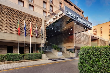 A hotel entrance with a modern facade, multiple flags, and a staircase leading to the entrance.. Sercotel Gran Hotel Zurbarán