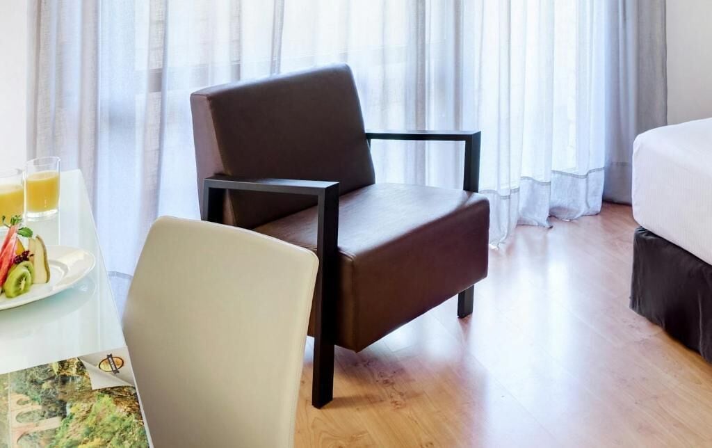 A hotel room with a bed, a chair, a table with a fruit plate, and a glass of orange juice.