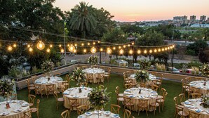 Un espacio de comedor al aire libre con mesas y sillas redondas, luces colgantes y un paisaje urbano de fondo.