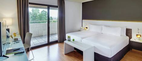 A hotel room with a bed, a desk with a laptop, a chair, a bedside table with a lamp, and a view of the outdoors.