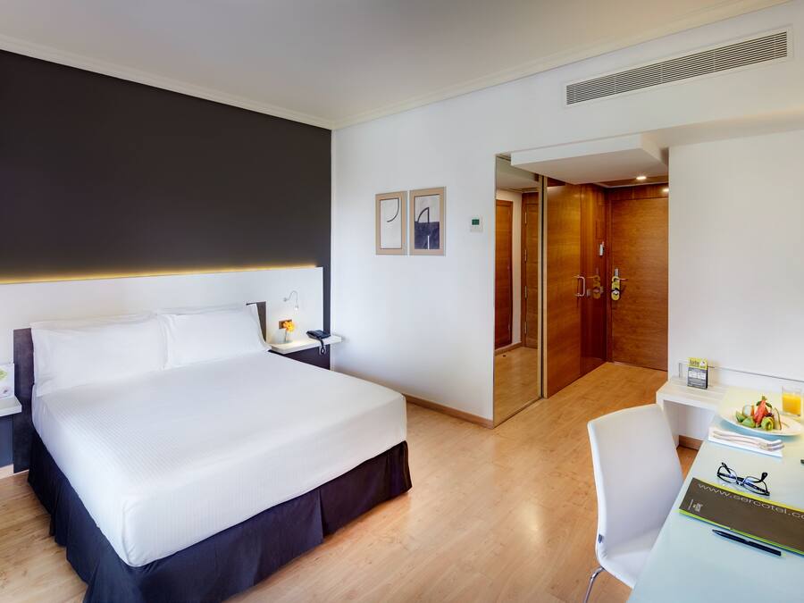 Habitación de hotel con una cama grande, un escritorio con una computadora, y una mesita con una bandeja de fruta y un vaso de jugo de naranja.