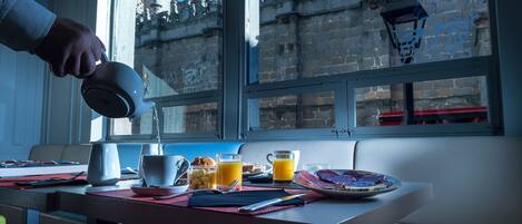 A breakfast table with a variety of food and drinks, a window with a view of a historic building, and a hand pouring a drink.