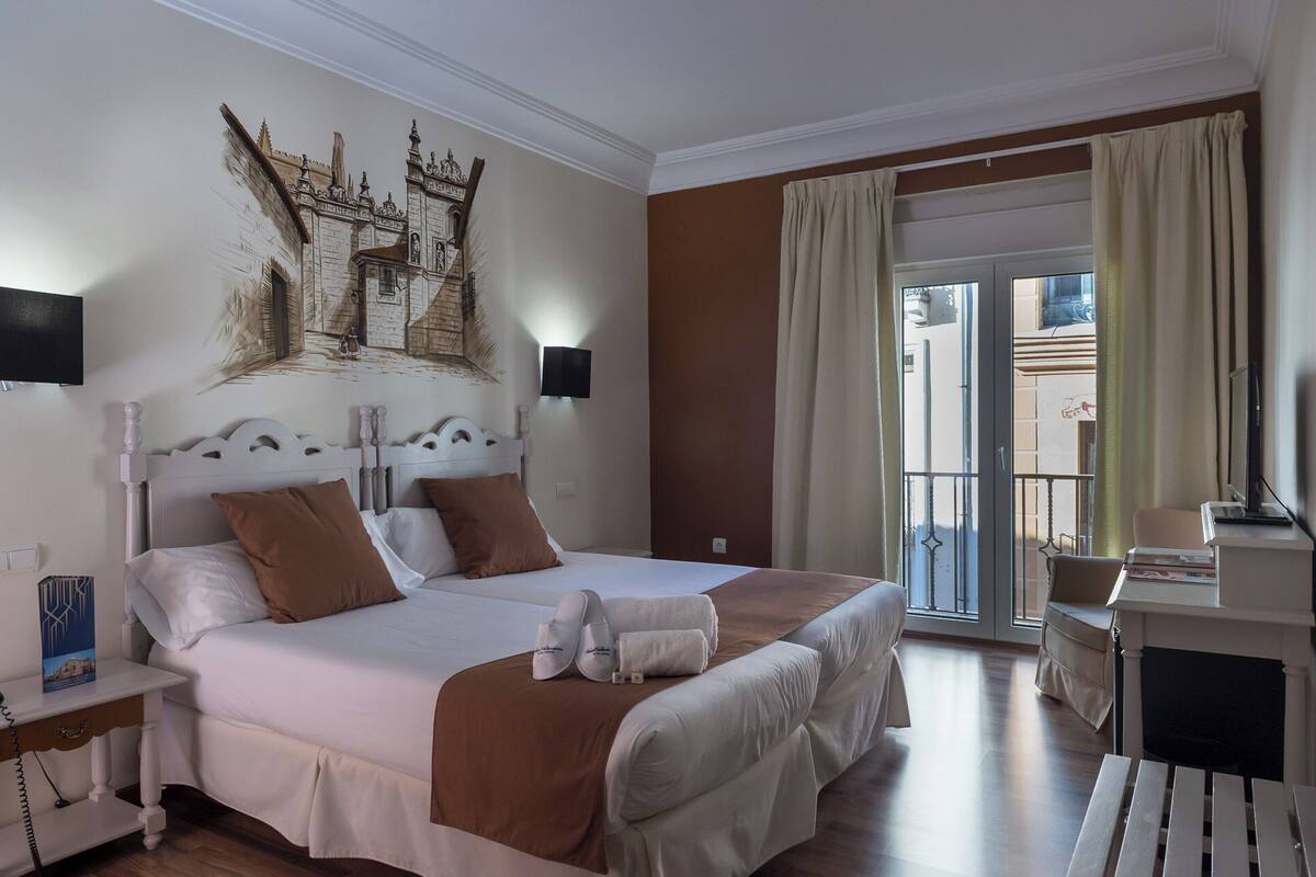 A hotel room with a large bed, a desk, a chair, a TV, and a balcony with curtains.