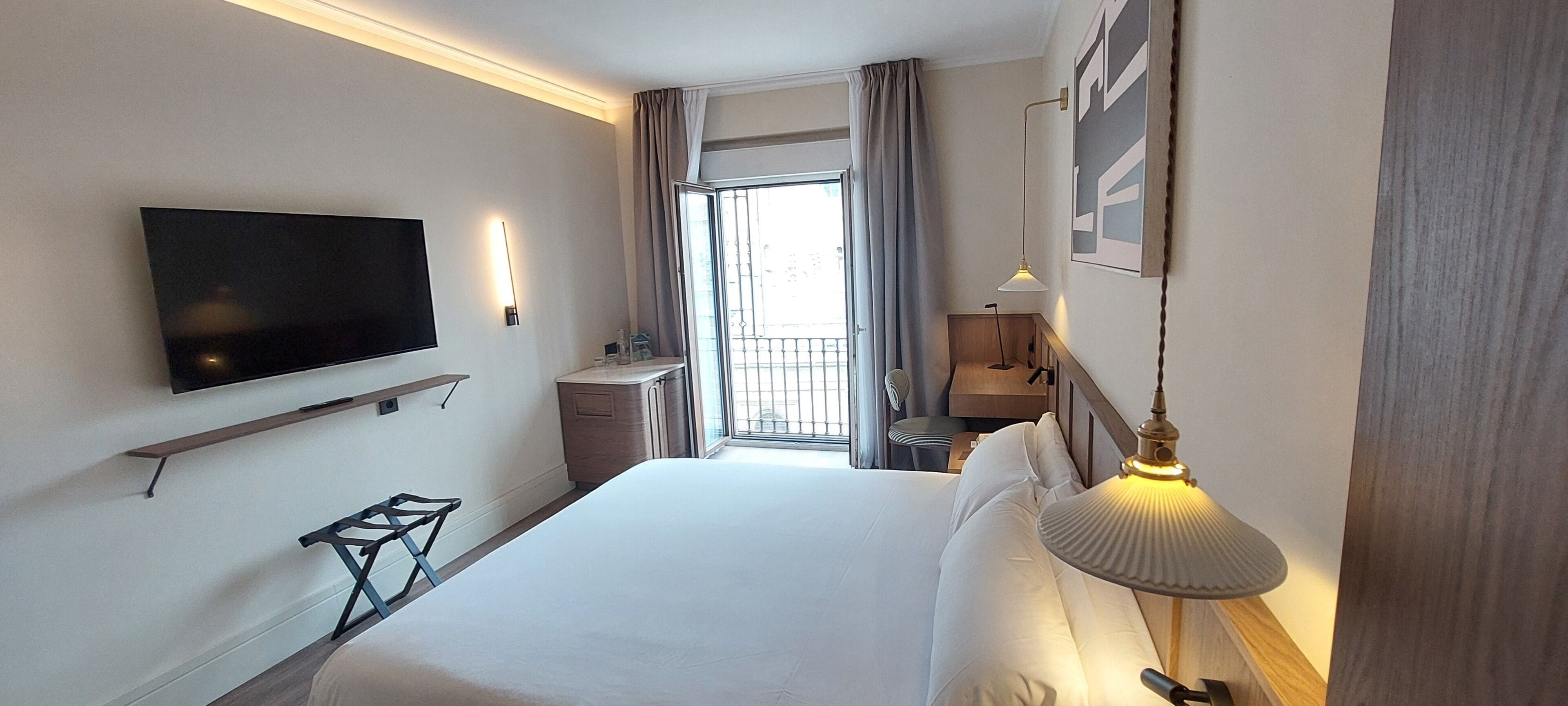 Habitación de hotel con cama, televisión montada en la pared, escritorio con silla, lámpara y ventana con cortinas.