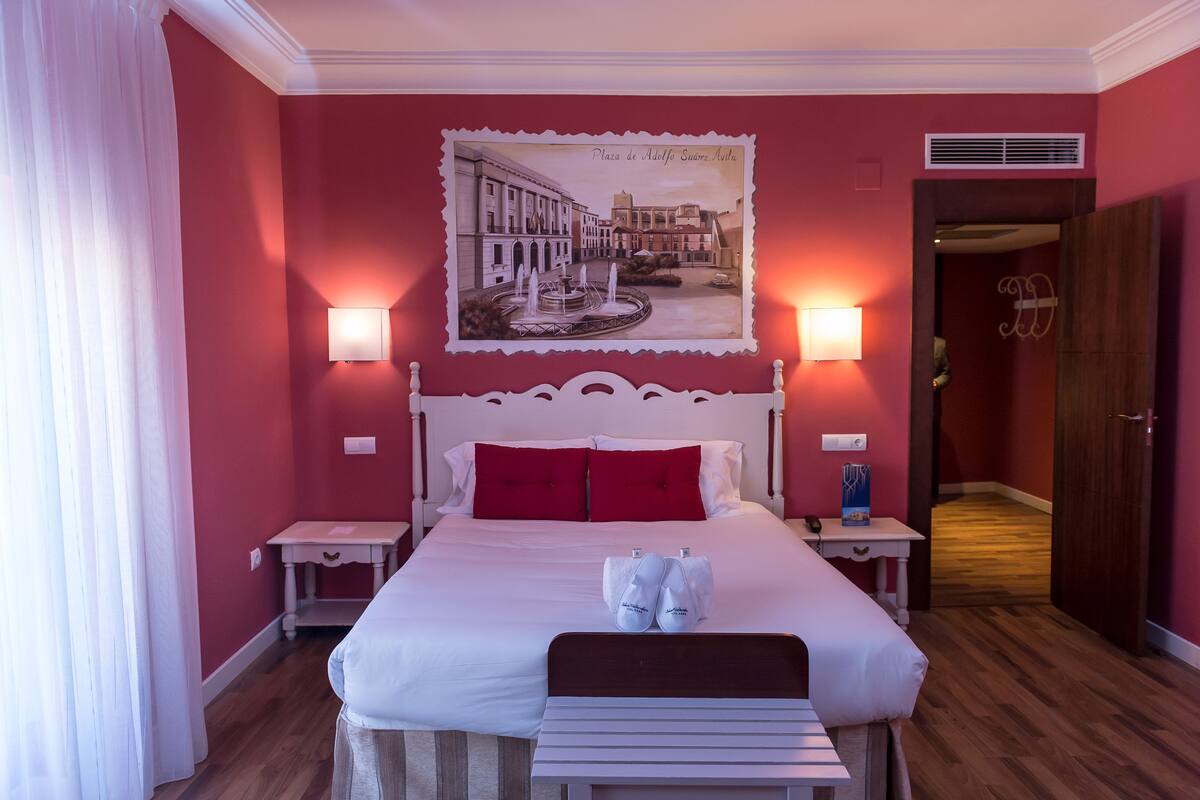 A hotel room with a large bed, red walls, a framed picture of a cityscape, two bedside tables with lamps, and a wooden bench.
