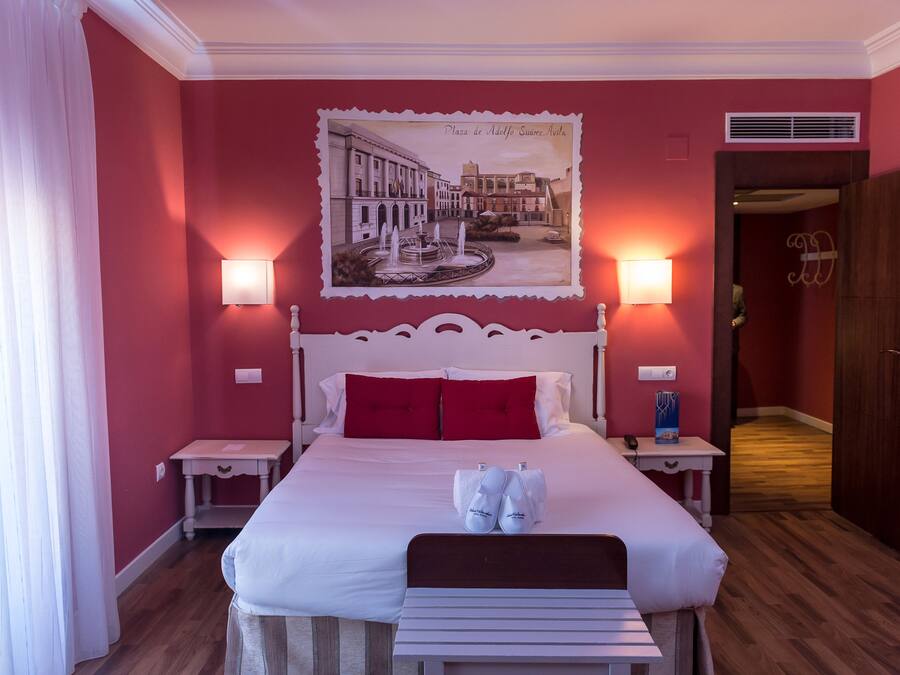 Una habitación de hotel con una cama grande, paredes rojas, un cuadro enmarcado de un paisaje urbano, dos mesitas de noche con lámparas y un banco de madera.