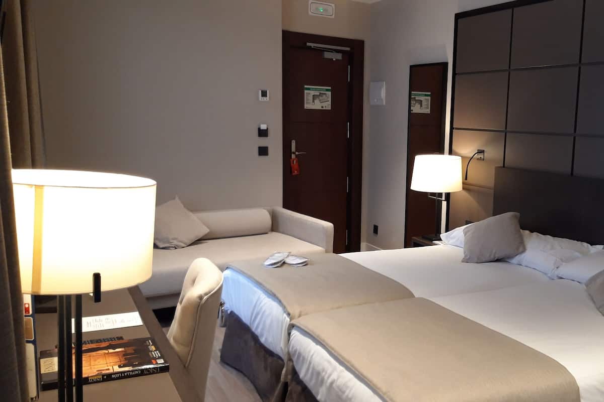 A hotel room with a large bed, a smaller sofa bed, a desk with a lamp, and a bedside table with a phone.