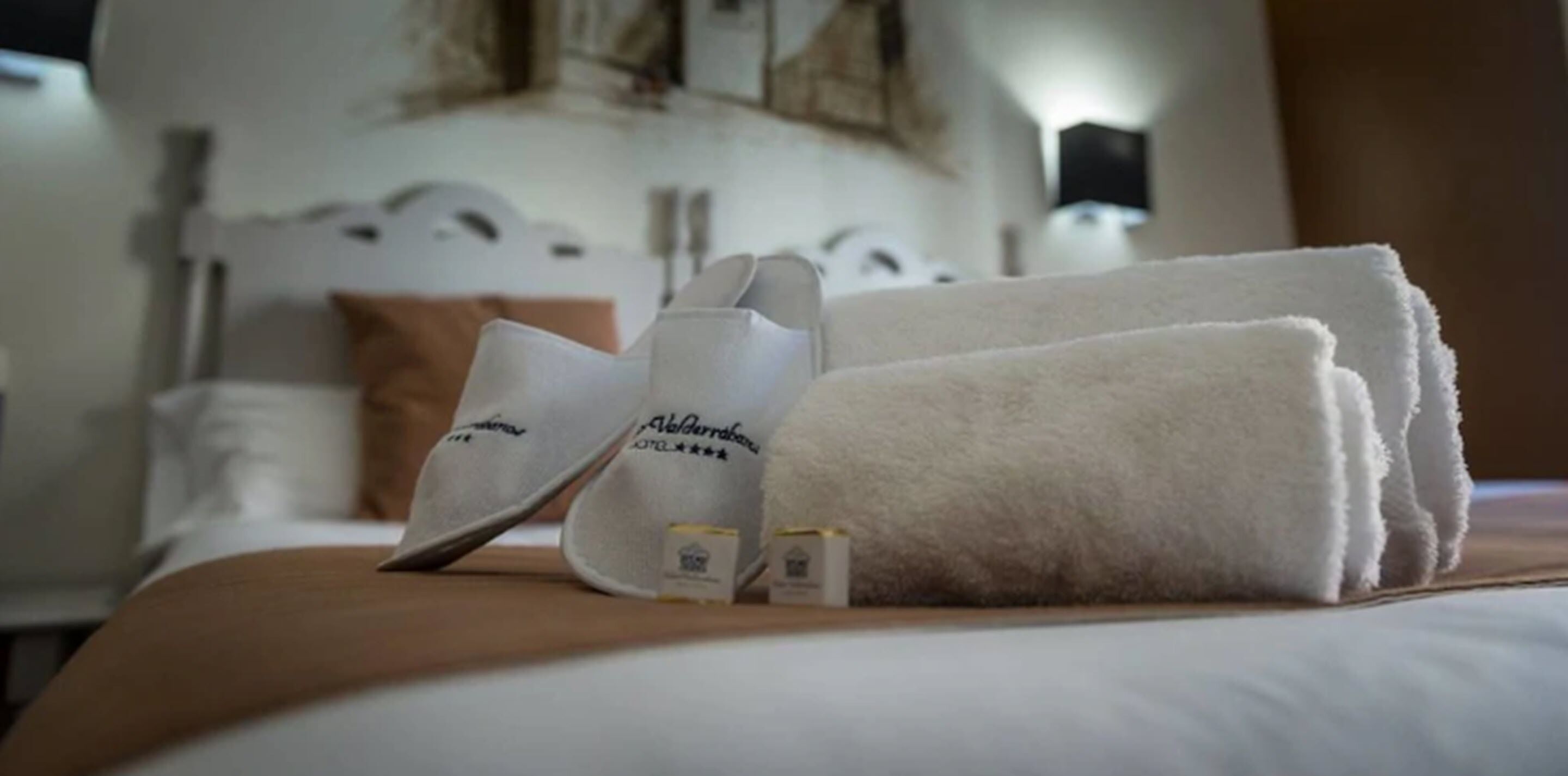 Una cama bien hecha con una toalla blanca doblada y dos pantuflas blancas con un logotipo.
