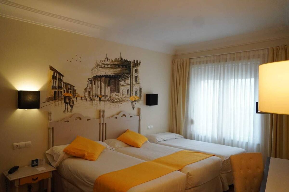 A hotel room with two beds, a mural on the wall, a desk with a lamp, and a window with curtains.