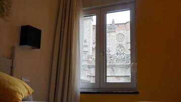 Una habitación con una cama, una ventana con cortinas, un televisor montado en la pared y vistas a un edificio de arquitectura detallada.