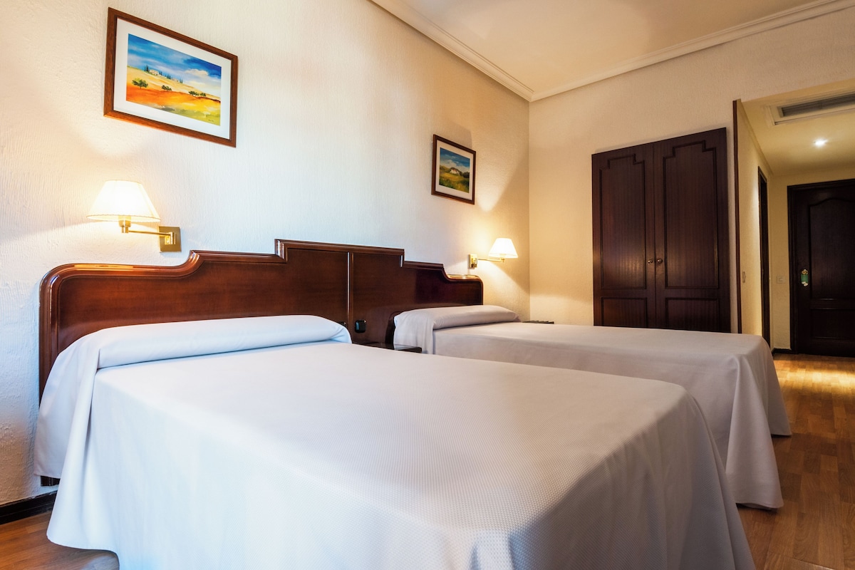 A hotel room with two single beds, a wooden headboard, wall-mounted lamps, and a door.