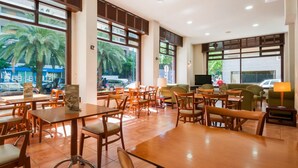 Un amplio espacio interior con varias mesas y sillas de madera, grandes ventanales que ofrecen vistas a palmeras y vehículos estacionados afuera.
