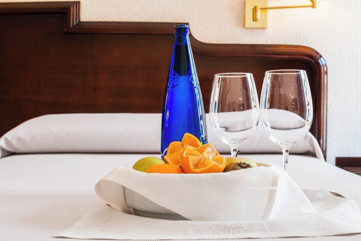 A neatly made bed with a white comforter, a blue bottle, two glasses, and a bowl of fruit.