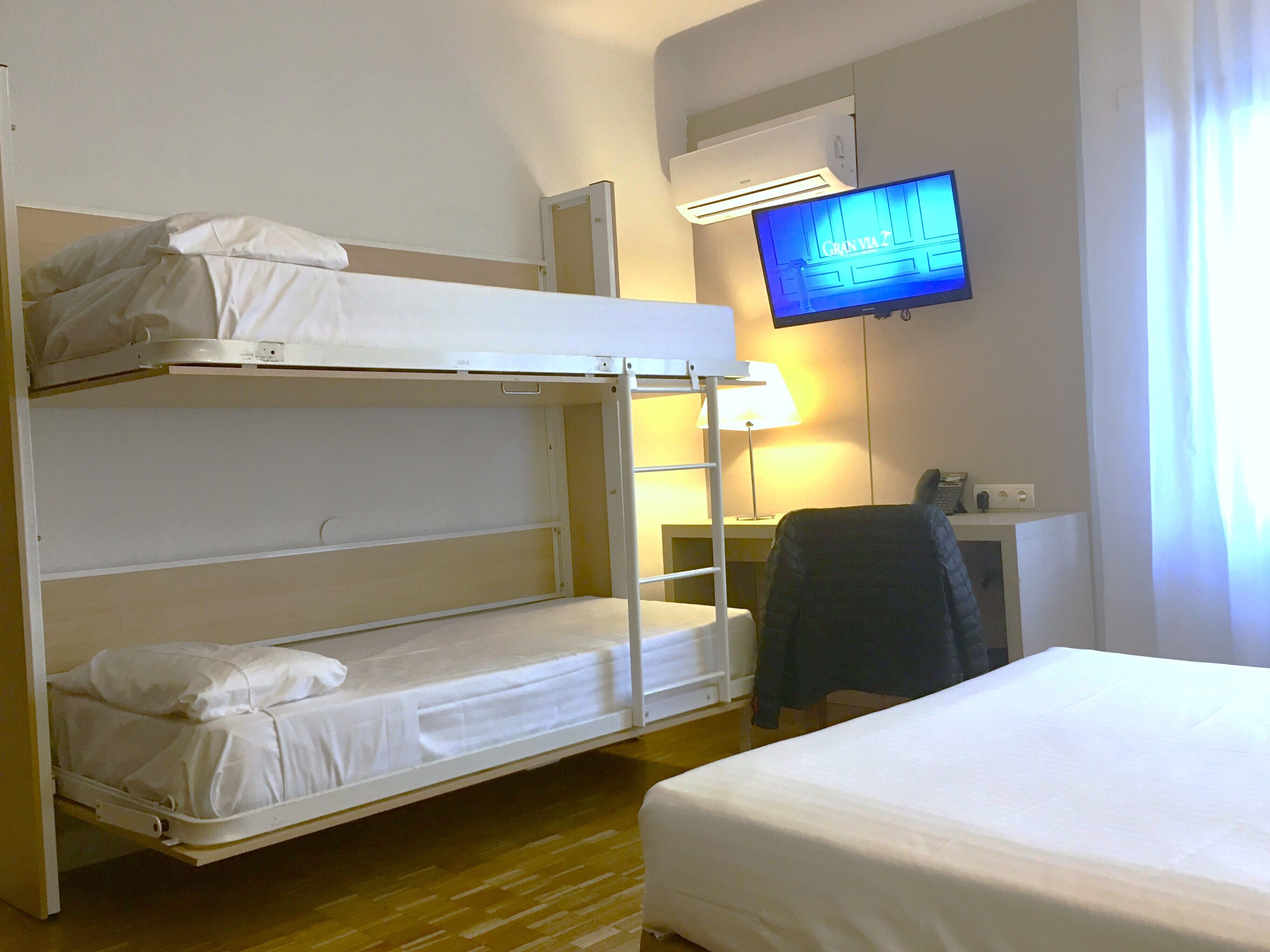 A hotel room with bunk beds, a desk with a chair, a television, and an air conditioning unit.