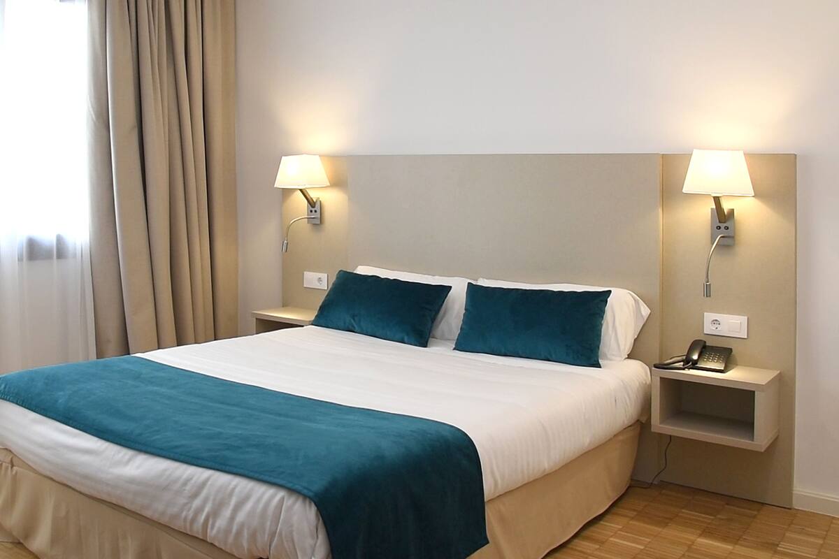 Una habitación de hotel con una cama grande, dos mesitas de noche con lámparas y un teléfono en una de ellas.
