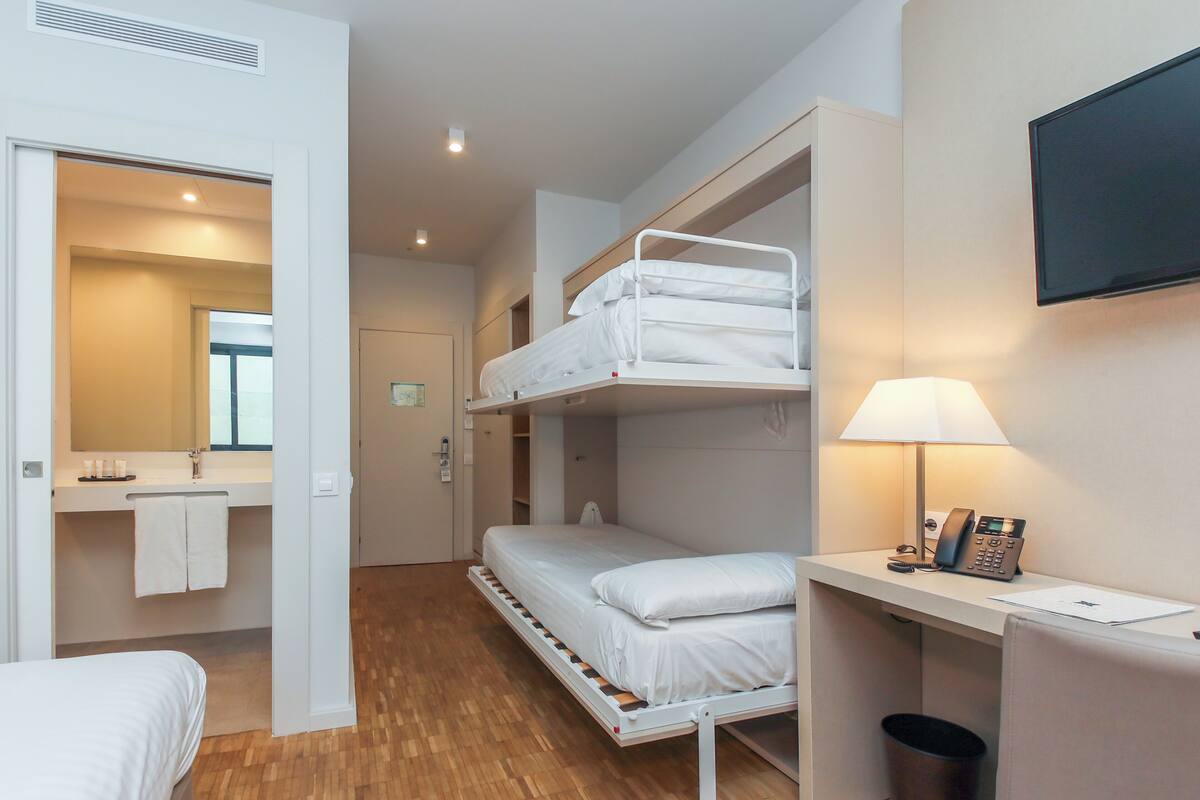 Una habitación de hotel compacta con una cama litera, un escritorio con lámpara, un teléfono y un espejo.