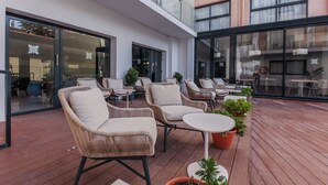 Een terras op het dak met rieten stoelen en tafels, grote ramen en potplanten.