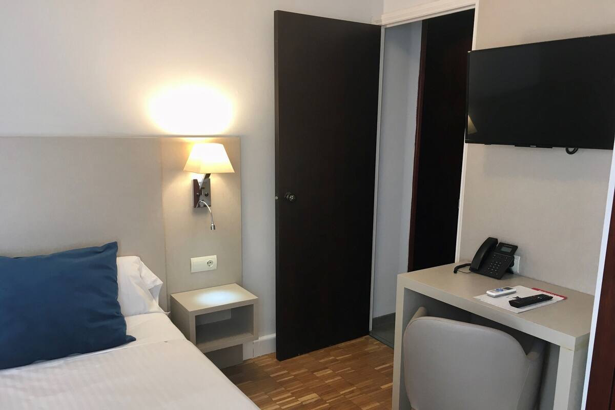 Una habitación de hotel con cama, escritorio, silla, lámpara de escritorio, teléfono y televisor.