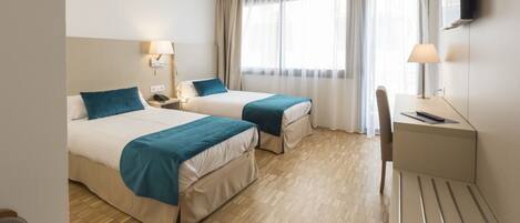 A hotel room with two beds, a desk, a chair, a TV, and a window with curtains.