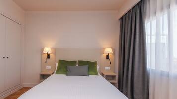 Una cama bien hecha con ropa de cama blanca, dos almohadas verdes y una almohada gris. Hay dos lámparas de noche a cada lado de la cama y un ventanal con cortinas grises.