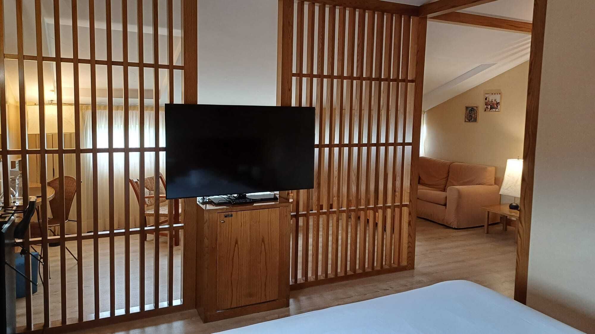 Ein Hotelzimmer mit Fernseher auf einem Holztisch, einem Bett, einer Couch und einer Essecke mit Stühlen und einem Tisch.