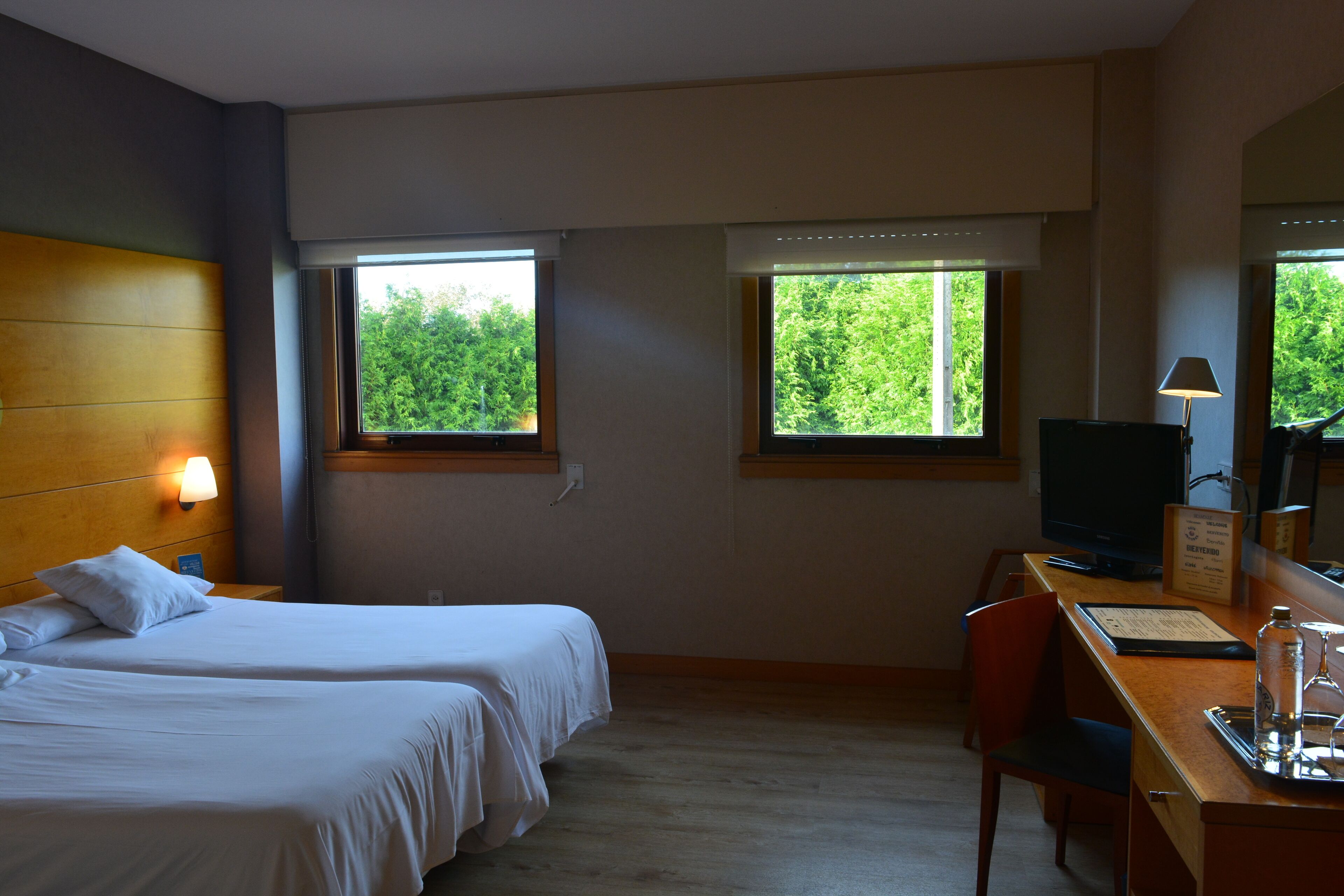 Ein Hotelzimmer mit zwei Betten, einem Schreibtisch mit Computer, einem Stuhl und Fenstern mit Blick auf Bäume.