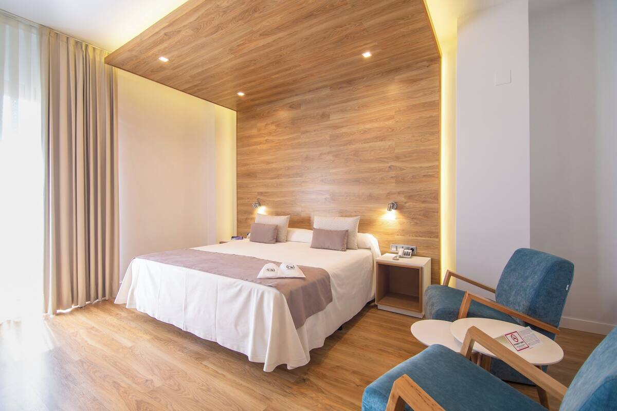 Una habitación de hotel moderna con techo de madera, una cama grande con colcha blanca y beige, dos mesitas de noche con lámparas, y un sillón azul con una mesita pequeña.