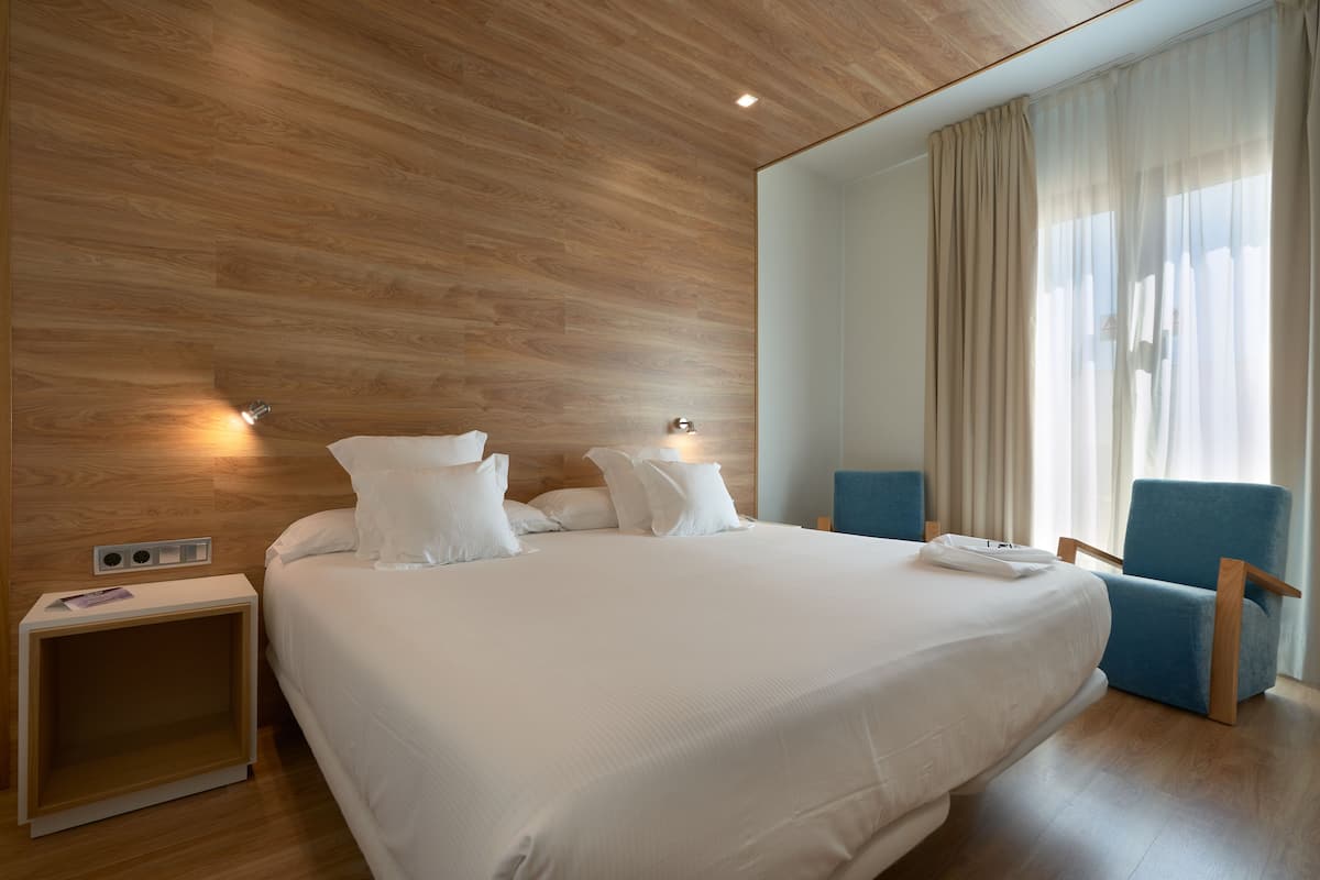 Una habitación de hotel moderna con una cama grande, una mesita de noche, una silla y un ventanal con cortinas.