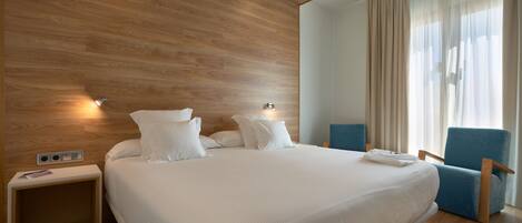 A modern hotel room with a large bed, a nightstand, a chair, and a window with curtains.
