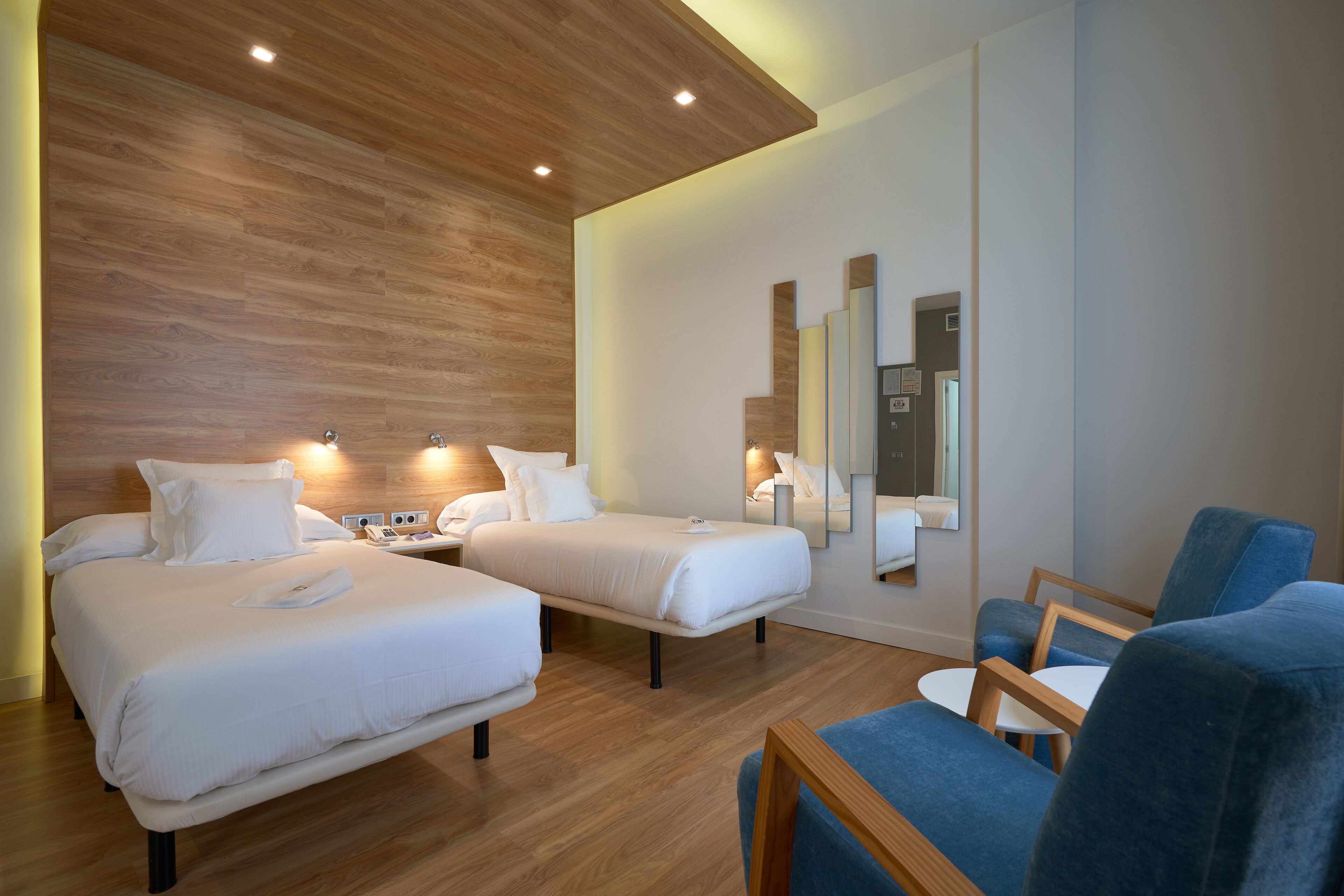 A hotel room with two beds, a blue armchair, a small table, and a wall-mounted mirror.