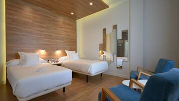 A hotel room with two beds, a blue armchair, a small table, and a wall-mounted mirror.