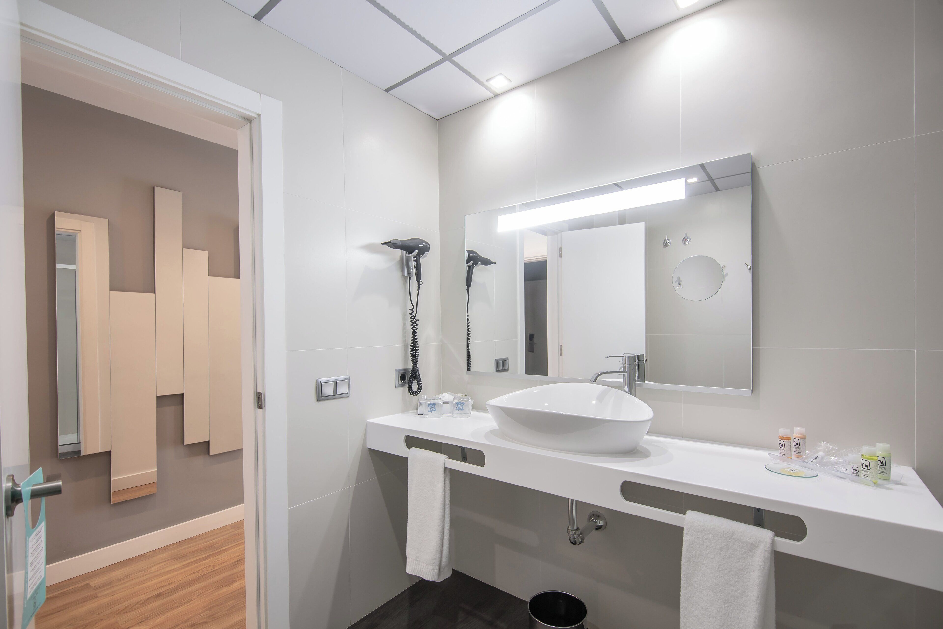 A modern bathroom with a large white sink, a mirror, and a hairdryer. There is a towel rack and a small trash can.