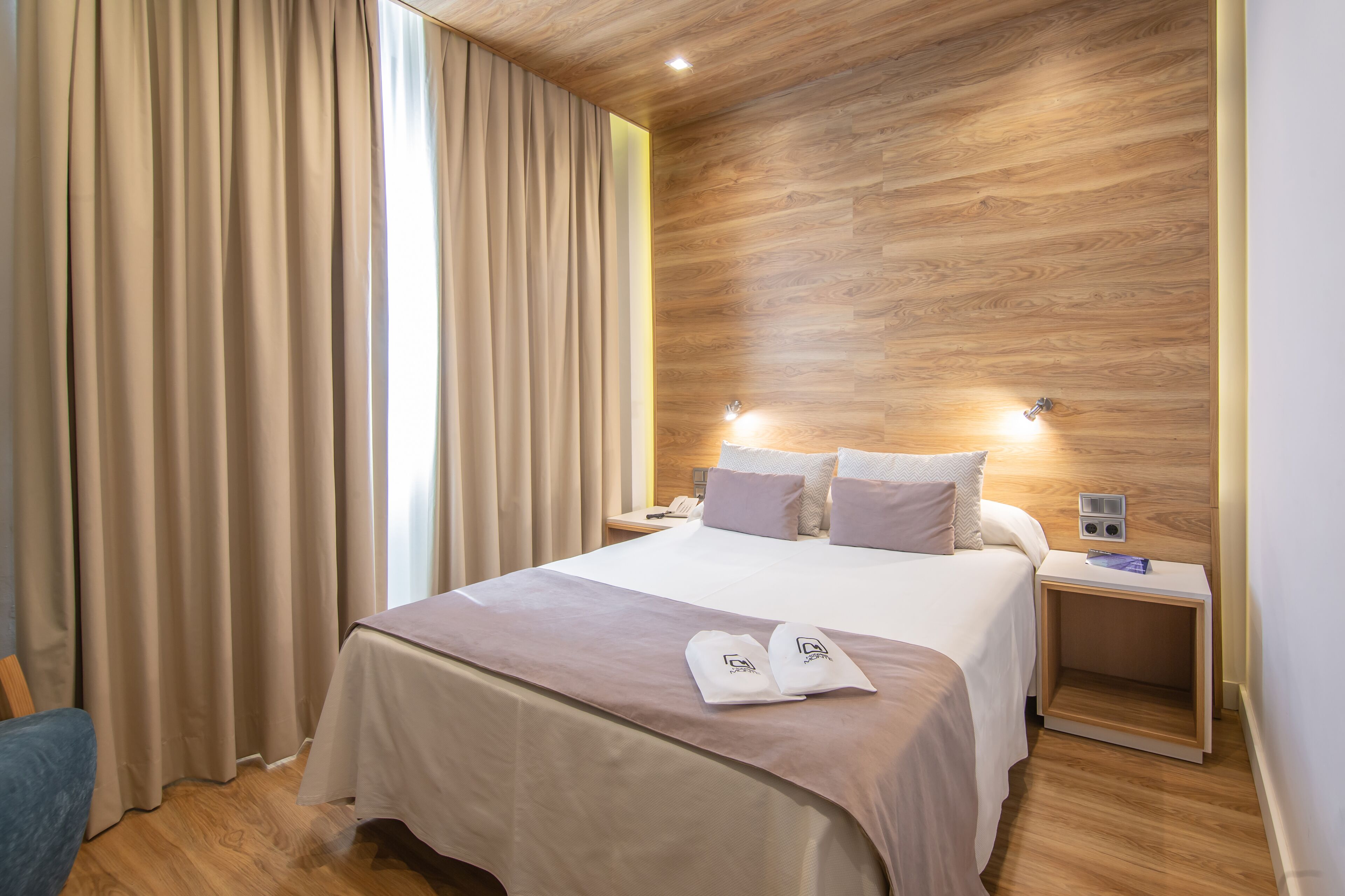 Quarto de hotel com parede de madeira, cama com roupa de cama branca, mesa de cabeceira com telefone e luminária, e janela com cortinas.