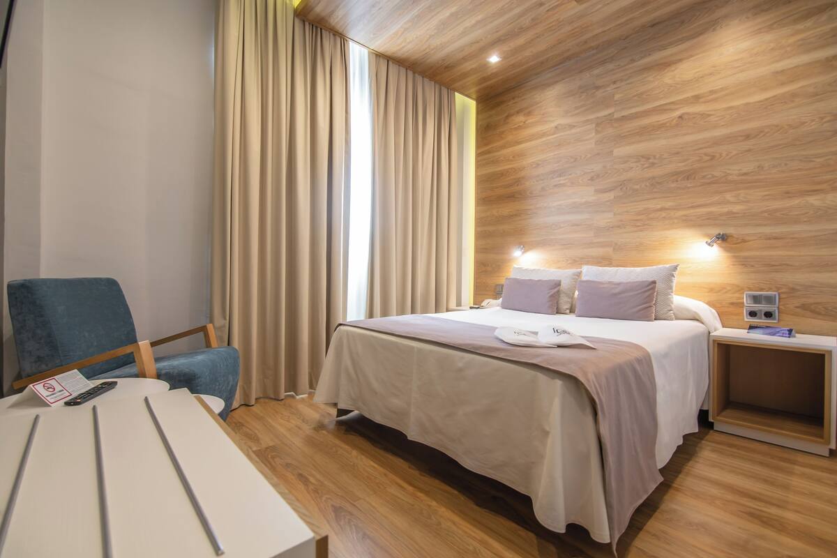 Una habitación de hotel moderna con techo de madera, una cama con ropa blanca, un sillón azul, una mesita de noche y una pequeña mesa auxiliar.