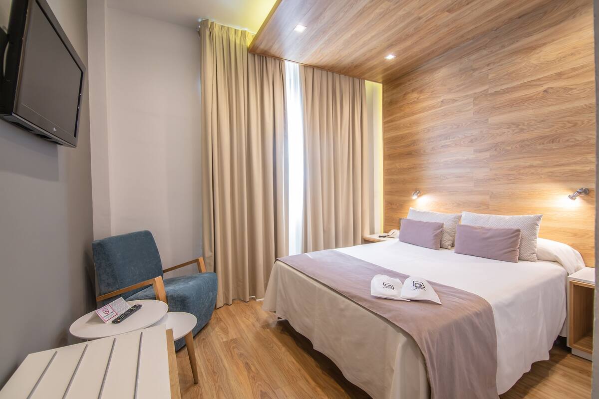 Una habitación de hotel con una cama, una silla, una mesita y un televisor colocado en la pared.