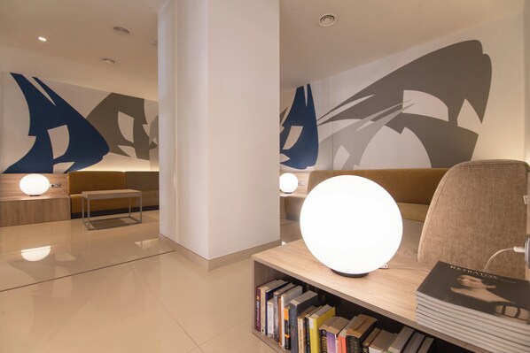 Un moderno vestíbulo de hotel con diseño minimalista, que presenta una zona de estar con un sofá y una mesita, una estantería con libros, y arte abstracto en la pared.
