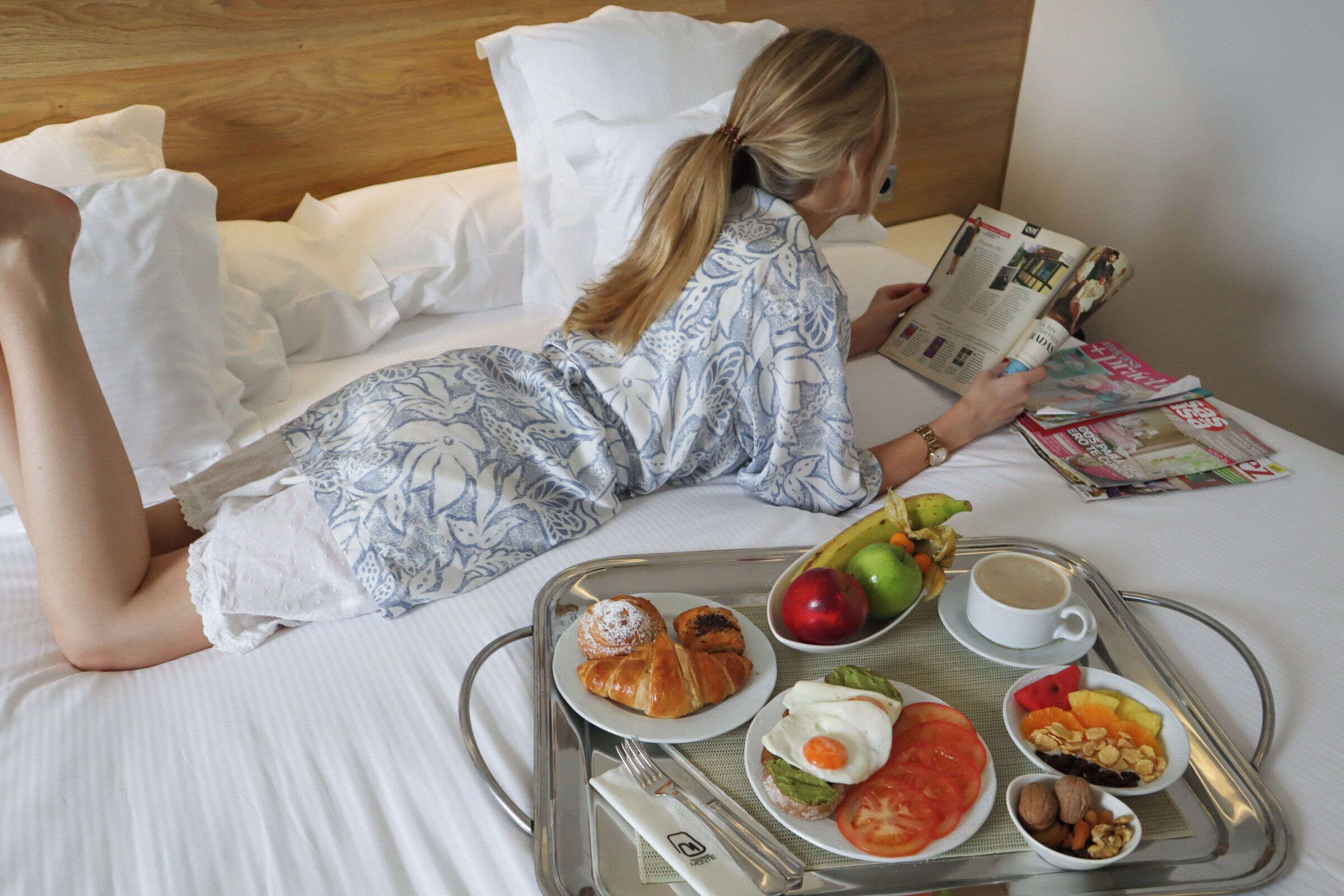 En person ligger i sängen och läser en tidning, och bredvid finns ett brickbord med frukost, inklusive croissanter, ägg, tomater och frukt.