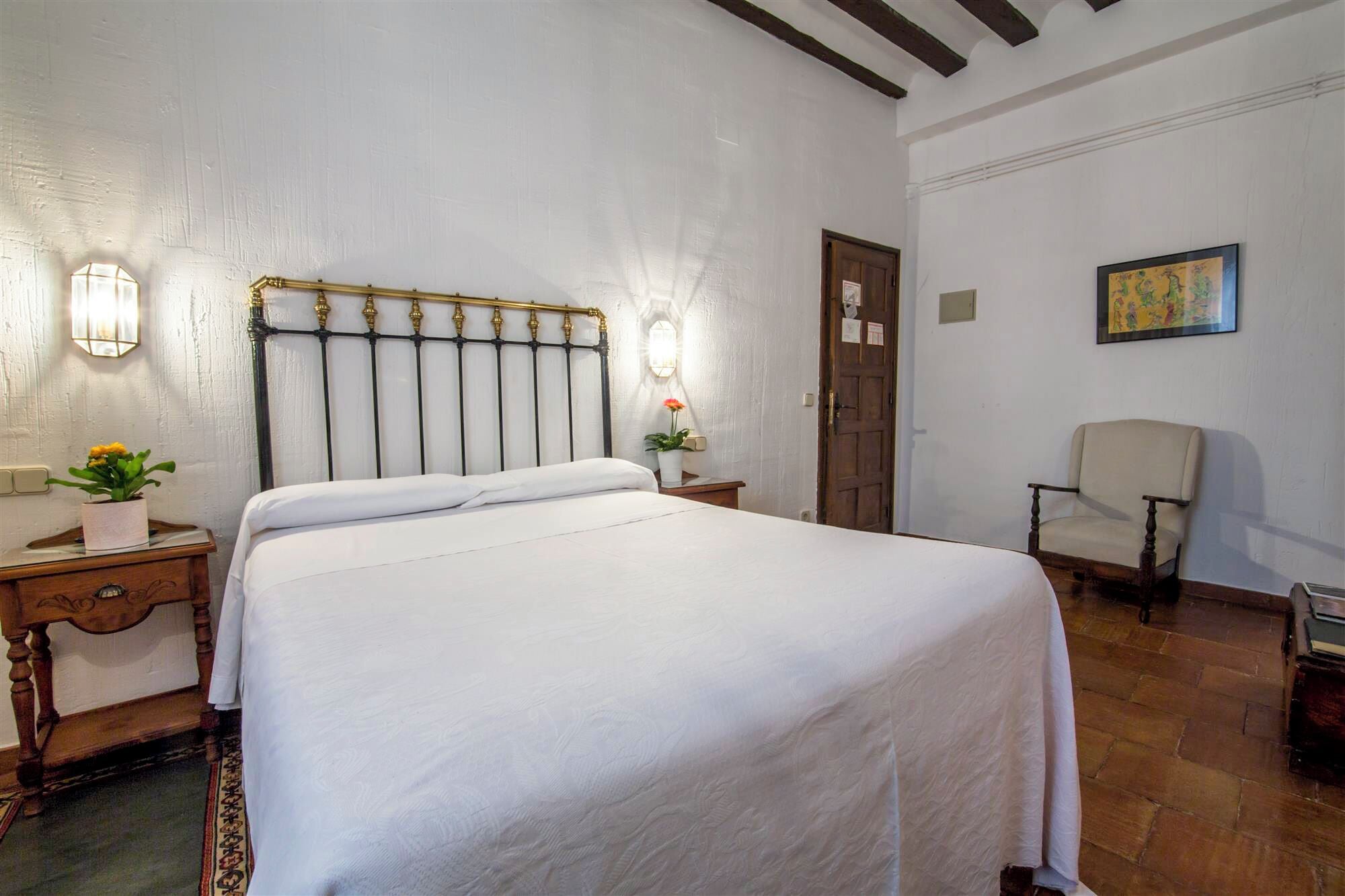 A neatly made bed in a room with a white wall and a wooden floor. There is a nightstand with a lamp and a flower vase, a chair, and a door with a picture on the wall.