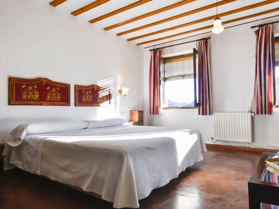 A bedroom with a large bed, wooden ceiling beams, a window with curtains, and a radiator.