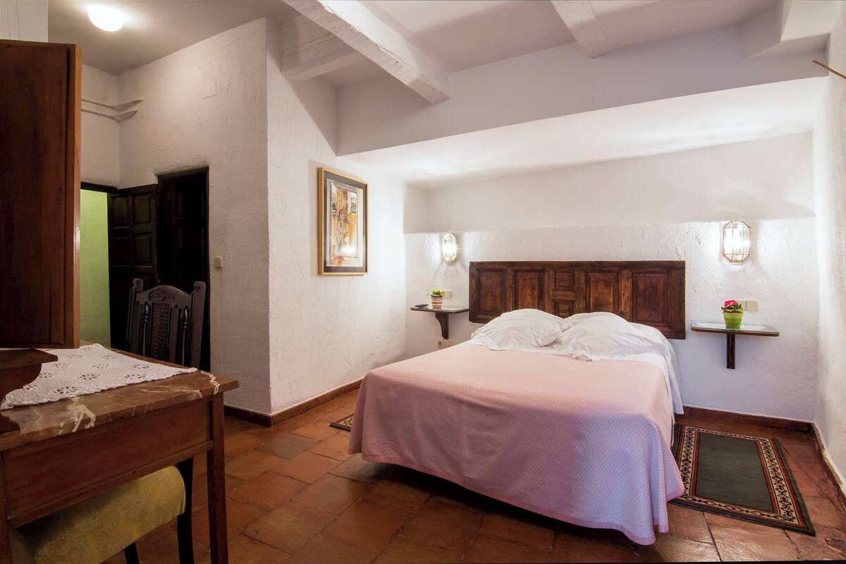 A bedroom with a wooden bed, a nightstand with a lamp, a chair, and a small table with a lace tablecloth.