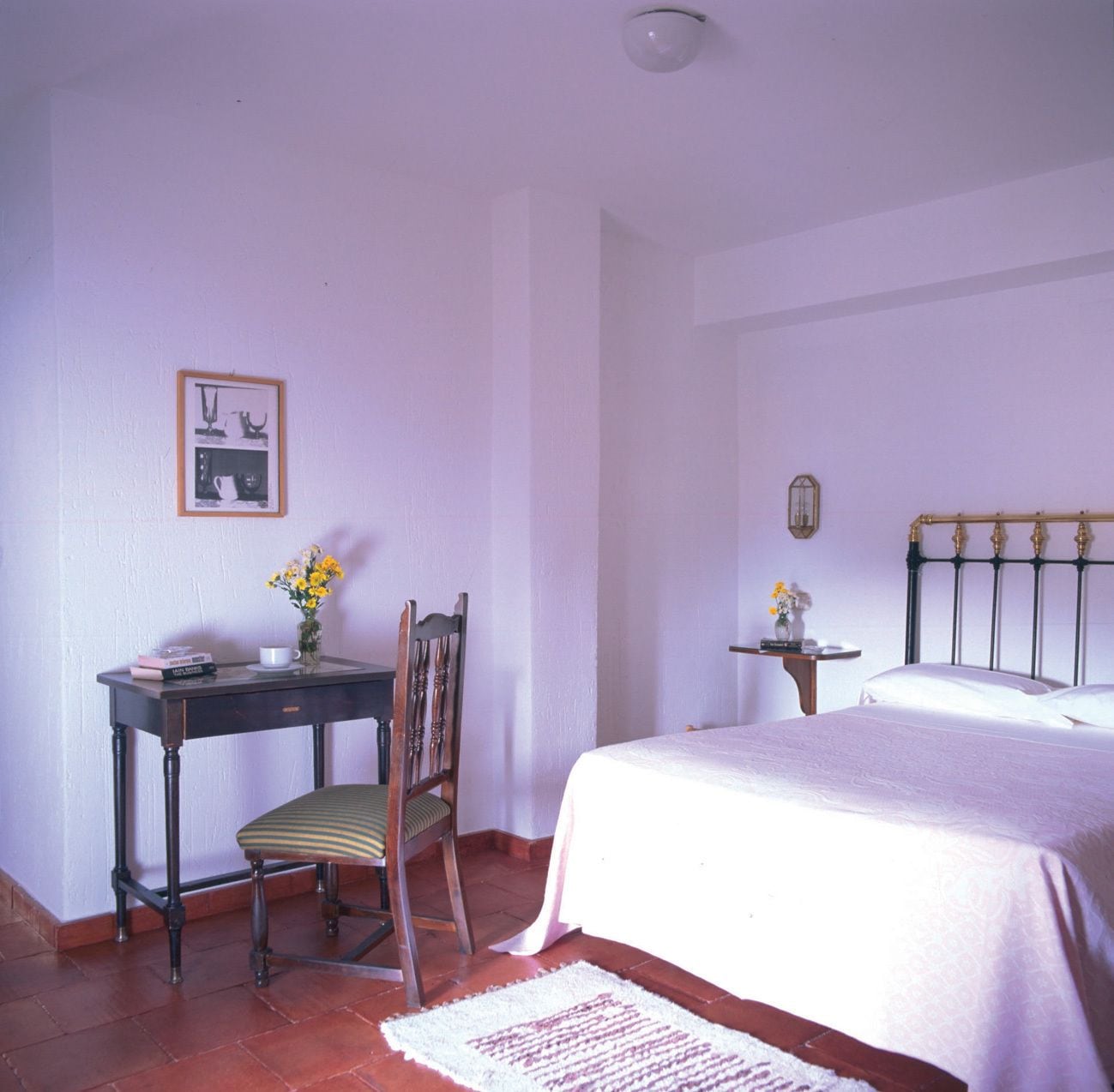 A bedroom with a bed, a desk with a chair, a small table with a vase, and a wall-mounted light fixture.